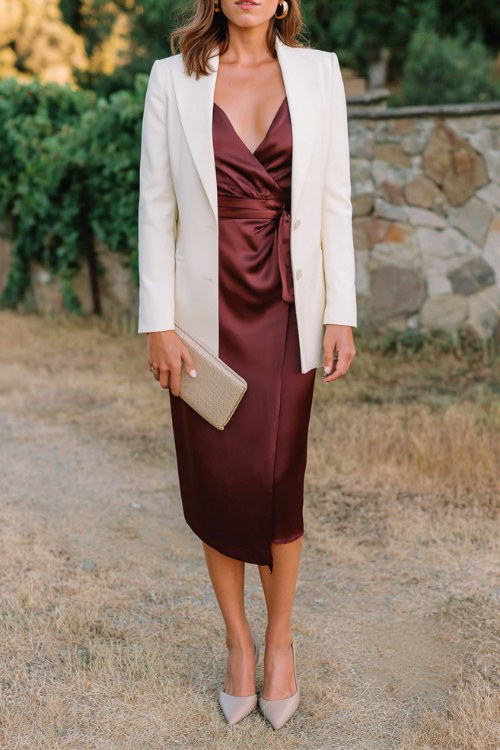 A woman wears a burgundy satin midi wrap dress with a belted waist, layered under a cream tailored blazer, paired with pointed-toe heels and a clutch