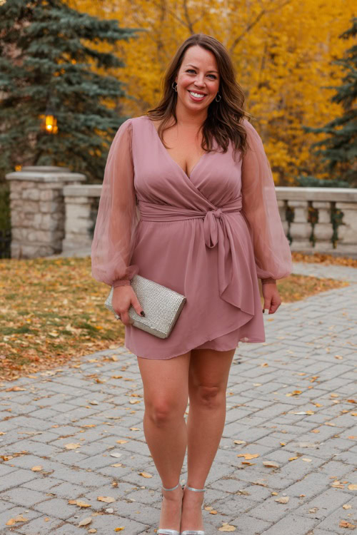 A plus-size woman wears a dusty rose wrap dress with long sheer sleeves and a tie waist, paired with silver block heels and a small clutch