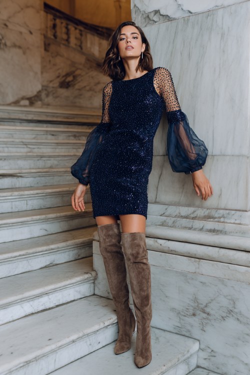A woman over 40 wears a midnight blue sequin sheath dress with sheer beaded sleeves, styled with tall heeled suede boots