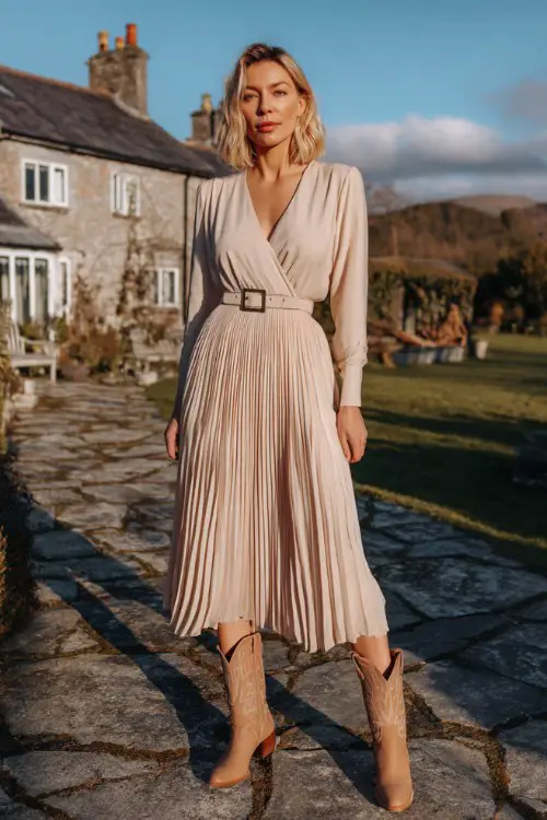 A woman over 30 wears a pleated midi dress in soft champagne with a V-neckline and a structured belt, paired with classic tan cowboy boots