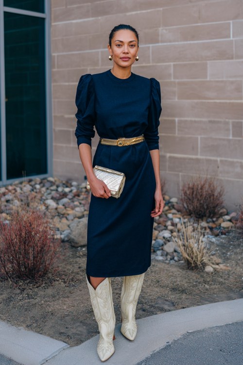 A woman in her 40s wears a navy blue wool-blend midi dress with structured shoulders and a gold belt, styled with embroidered ivory cowboy boots