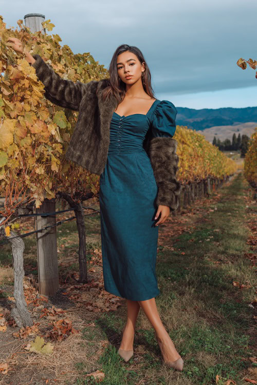 A woman wears a deep teal midi dress with puff sleeves, paired with a cropped faux fur jacket and heeled booties