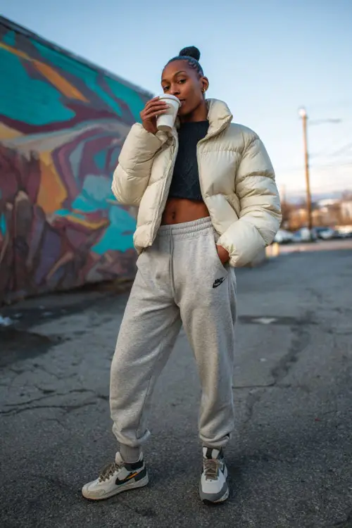 Black woman in a cropped puffer jacket, jogger pants, and sneakers, sipping coffee while standing in front of a mural on a chilly urban morning