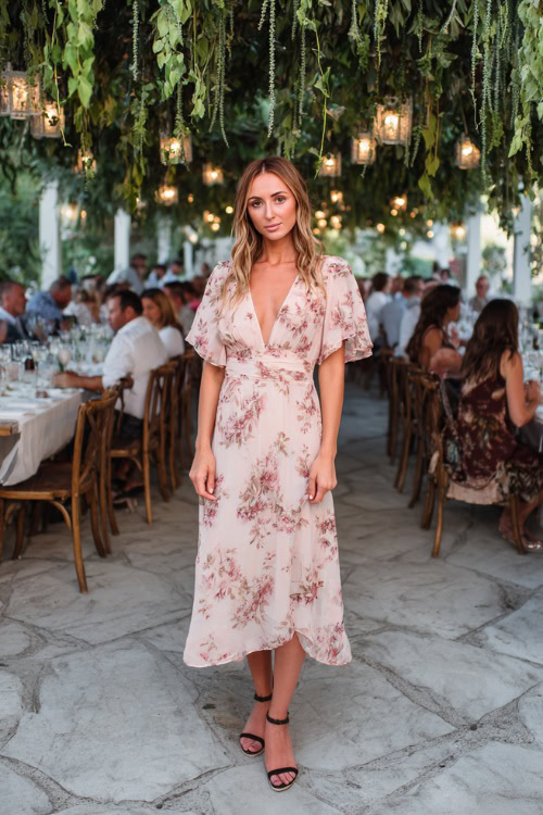 A woman wearing a blush pink floral flowy midi dress with flutter sleeves and wedge sandals, standing beneath a canopy of hanging greenery at an outdoor summer wedding reception