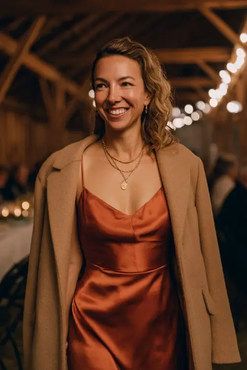 A woman over 35 wears a rust-colored satin dress, light camel coat draped over her shoulders, and gold jewelry
