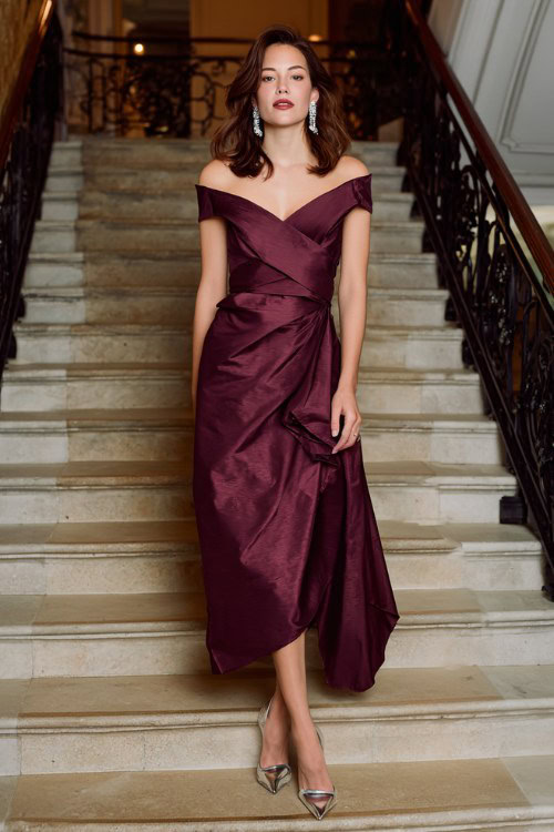 A woman wears a rich plum wrap-effect gown with an asymmetric neckline and satin texture, paired with chrome heels and chandelier earrings