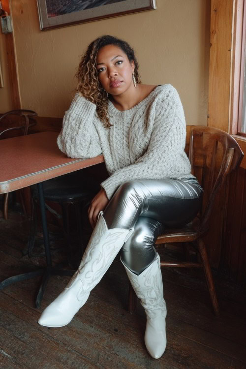 A curvy woman wears shiny faux-leather leggings with white tall cowboy boots and an oversized cable-knit sweater