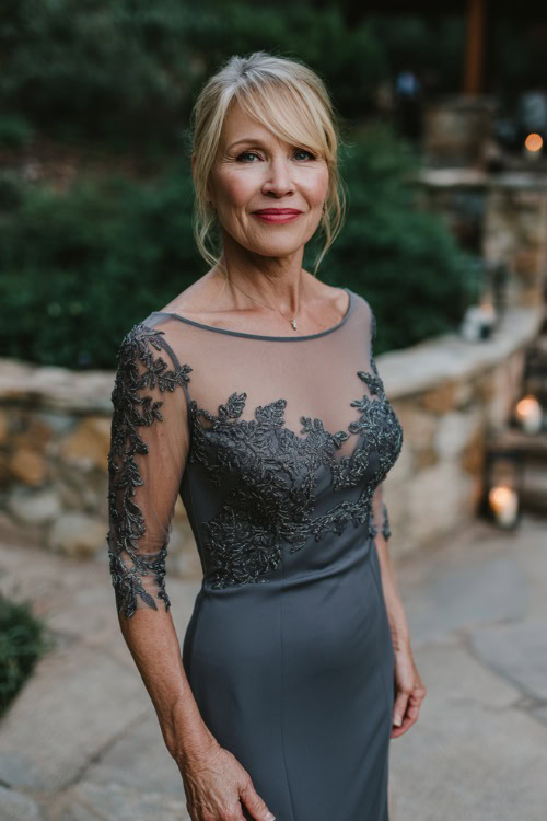 A woman over 50 wearing a charcoal gray embellished gown with illusion sleeves and a subtle mermaid silhouette, posing near a candlelit pathway at a twilight garden wedding