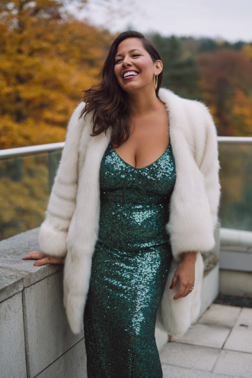 A curvy woman wears a long emerald sequin gown with a white faux fur coat