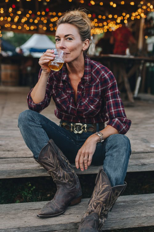 A woman over 40 wears classic denim jeans, a maroon and navy plaid flannel tucked in with a statement belt buckle, and dark leather cowboy boots