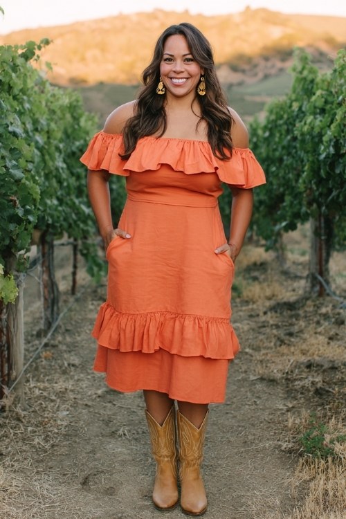 A curvy woman wears a bright coral off-shoulder midi dress with ruffled layers, styled with tan cowboy boots and gold hoop earrings