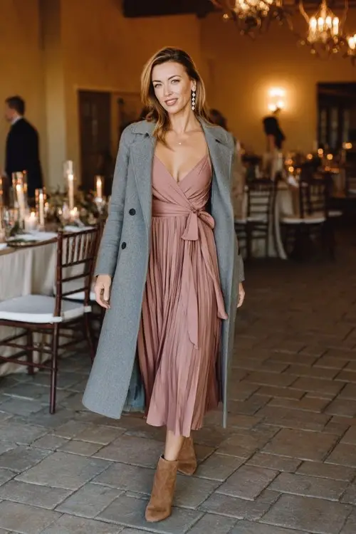 A woman wears a dusty rose midi dress with subtle pleating, layered under a soft grey wrap coat with a tie belt, suede ankle boots, and pearl earrings