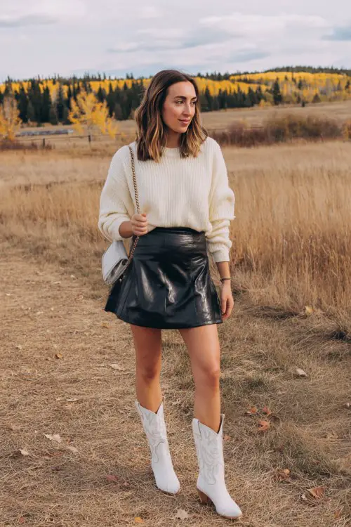 A woman wears a black faux-leather skater skirt with a cream knit sweater tucked in, paired with white cowboy boots and a crossbody bag