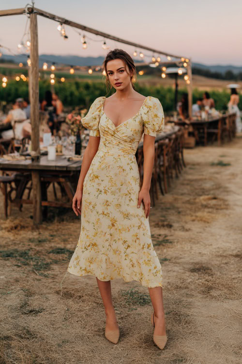 A woman wearing a pastel yellow floral midi dress with short puff sleeves and wedge heels, posing under hanging string lights at a vineyard wedding reception