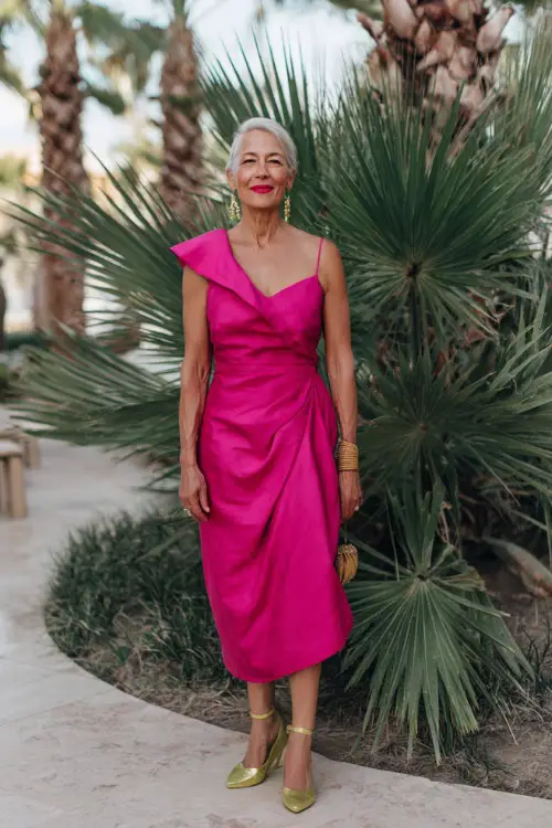 A woman over 50 wearing a bold fuchsia cocktail dress with asymmetrical neckline and gold heels, standing under tropical palms at a beachfront summer wedding reception
