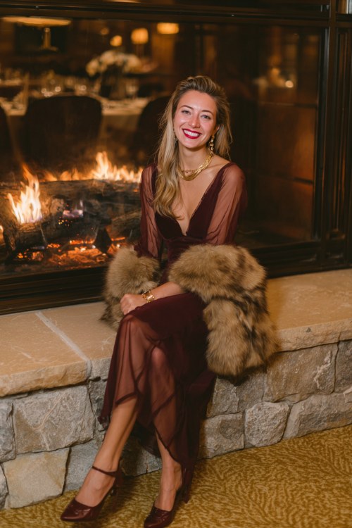 A woman wears a deep wine-colored dress with sheer sleeves and a faux fur stole, gold accessories, and brown pumps