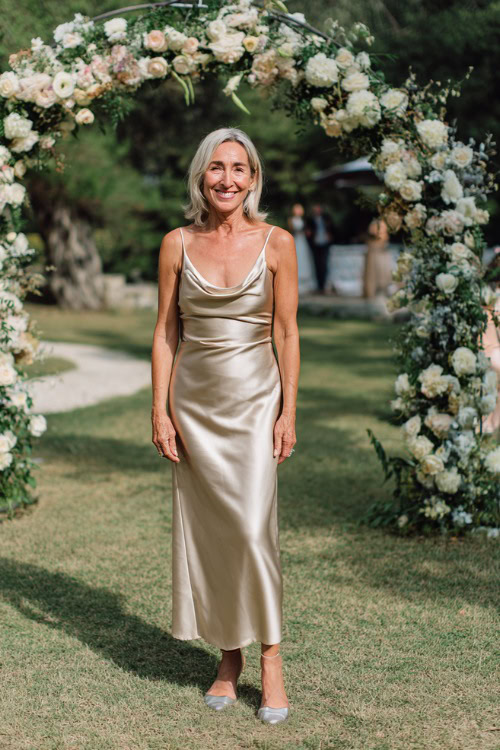 A woman over 50 wearing a floor-length champagne satin gown with a cowl neckline and silver heels, standing under a floral arch at a high-end garden wedding