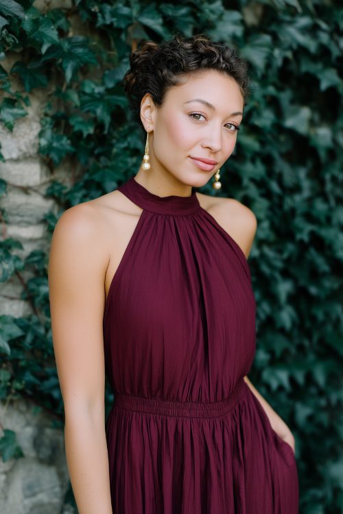 A woman in a deep wine-colored midi dress with a high neckline, pleated skirt, and pearl drop earrings