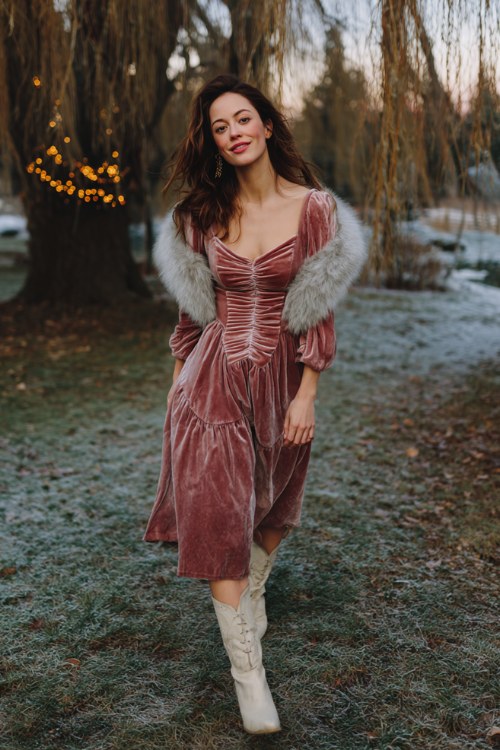 A woman wears a dusty rose velvet dress with a ruched bodice and sheer sleeves, topped with a soft grey faux fur collar and ivory suede boots