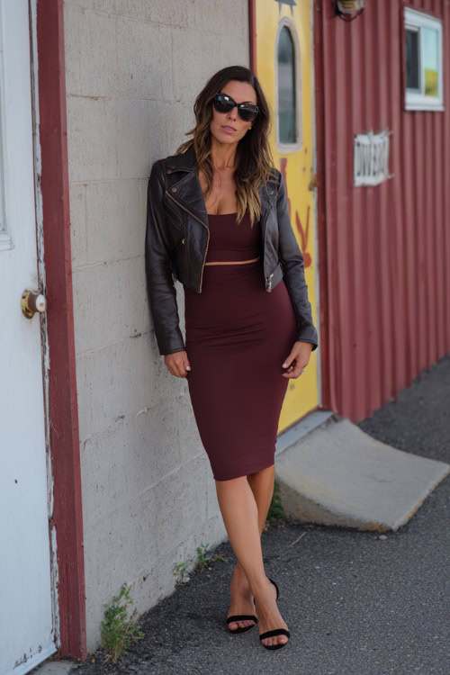A woman over 40 in a burgundy bodycon midi dress with a cropped leather jacket and black heels, posing near a casual fall bar entrance
