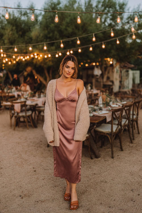 A woman wears a dusty rose midi slip dress with a soft knit cardigan layered on top, styled with block heels, standing in a backyard wedding setting with string lights and rustic tables