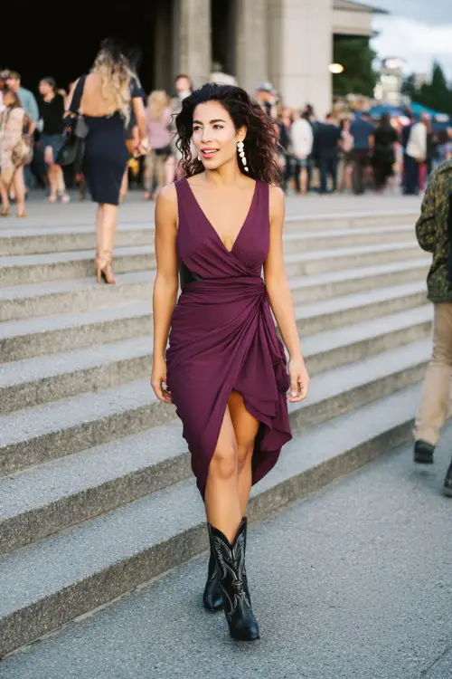A woman wears an elegant plum wrap dress with black heeled cowboy boots and pearl earrings