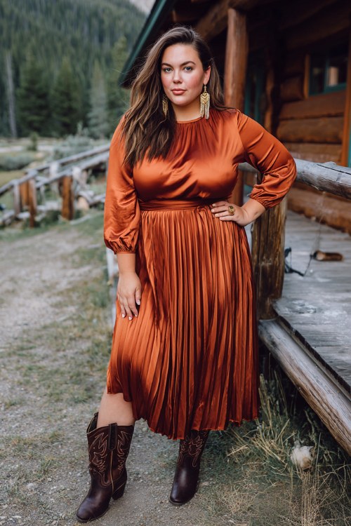 A plus-size woman wears a copper satin pleated dress with long sleeves, paired with tall dark brown cowboy boots and gold drop earrings