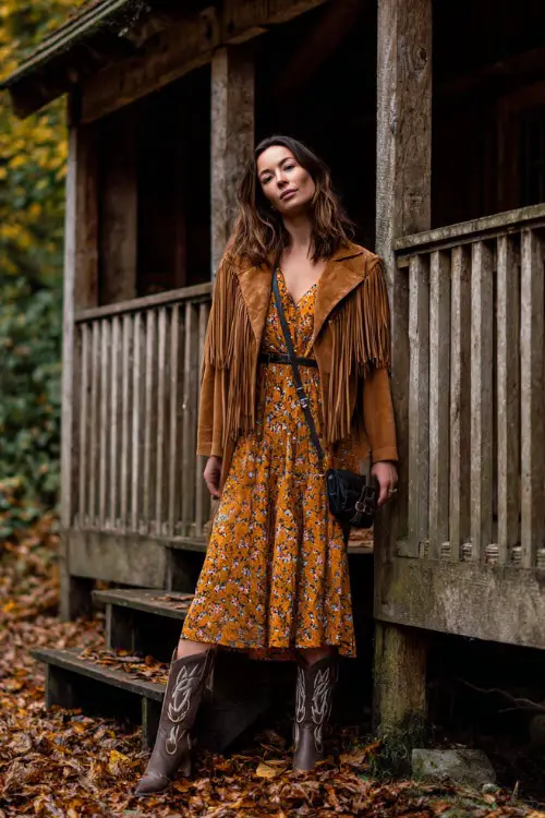 A woman wears a floral long sleeve midi dress with a suede fringe jacket, styled with cowboy boots and a crossbody bag, standing in a rustic outdoor stage surrounded by autumn leaves