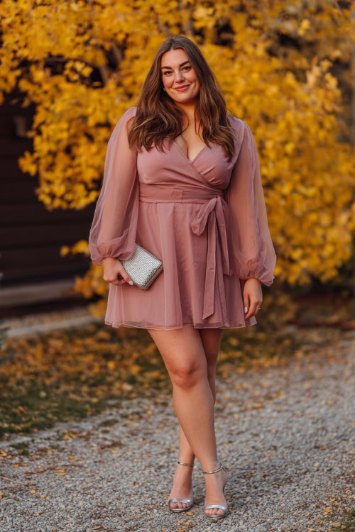A plus-size woman wears a dusty rose wrap dress with long sheer sleeves and a tie waist, paired with silver block heels and a small clutch