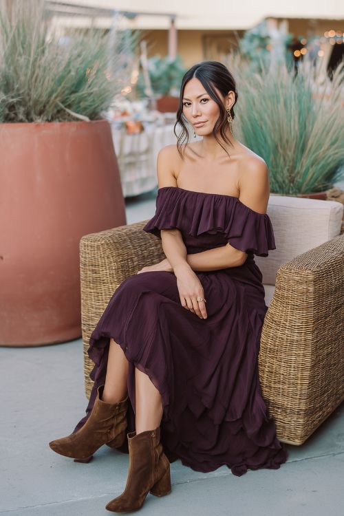 A woman wears a deep plum off-the-shoulder dress with a flowy hem and heeled brown boots