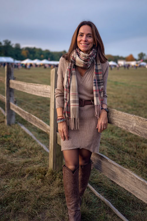 A woman over 50 wears a belted sweater dress with opaque tights, paired with tall western boots and a plaid scarf, standing near a wooden fence at a fall concert field