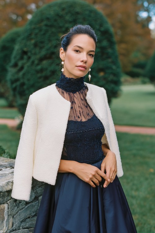 A woman wears a navy high-neck gown with sheer beaded sleeves and a flowy skirt, styled with a white boucle cropped jacket