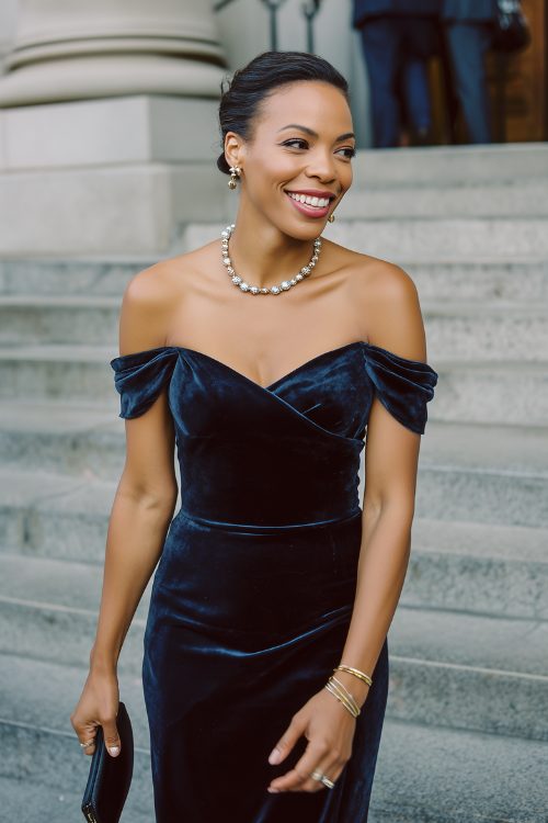 A black woman in a navy velvet gown with off-the-shoulder sleeves and pearl accessories