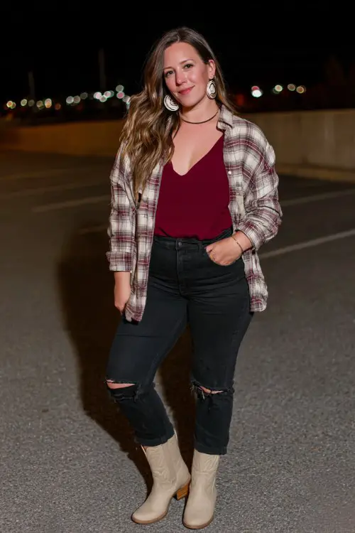 A curvy woman wears ripped black jeans with a fitted burgundy top layered under an oversized flannel, styled with beige cowboy boots