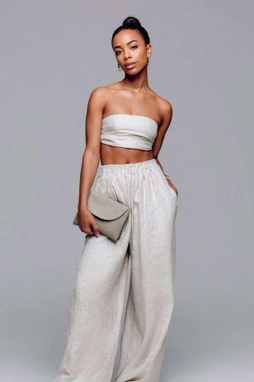 A woman in a lightweight linen two-piece set with a bandeau top and flowy pants, paired with slides and a clutch, styled for a cool downtown vibe
