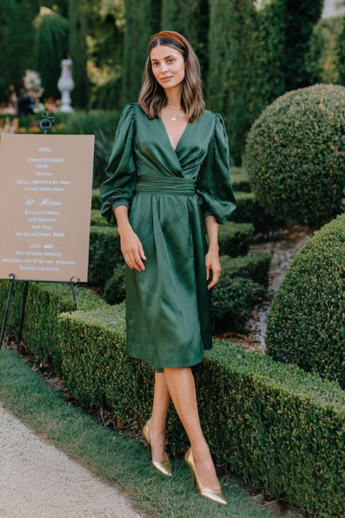 A petite woman wears a forest-green wrap midi with balloon sleeves and a waist sash, paired with gold slingback pumps