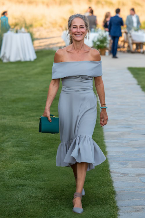 A woman over 50 wearing a soft grey off-the-shoulder cocktail dress with ruffled hem and clutch bag, walking across a lawn set up for an intimate summer wedding