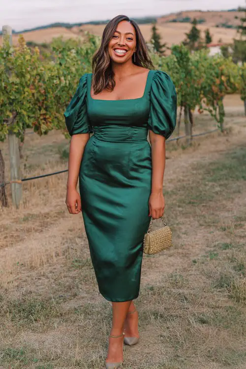 A curvy woman wears an emerald-green satin midi dress with a square neckline and gathered sleeves, paired with classic pumps