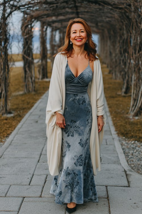 A woman wears a slate blue fit-and-flare dress with velvet floral overlay and bell sleeves, paired with a long ivory cape wrap
