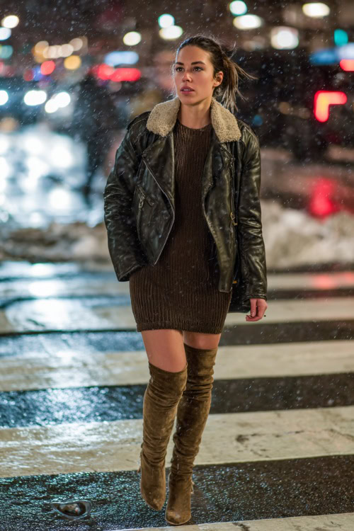 Trendy woman in a faux leather jacket, fitted knit dress, and thigh-high suede boots, walking through a snowy city crosswalk at night with lights reflecting off wet streets