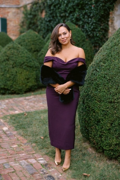 A curvy woman wears a deep purple off-shoulder gown with a black fur stole draped elegantly across her arms