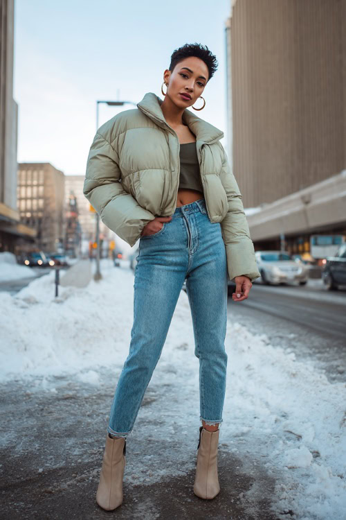 Confident woman in a cropped puffer jacket, high-waisted skinny jeans, heeled ankle boots, and hoop earrings, posing on a snowy city street with attitude