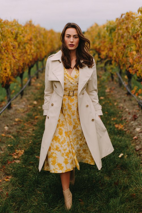 A woman wears a mustard yellow floral tea-length dress with a belted cream trench coat