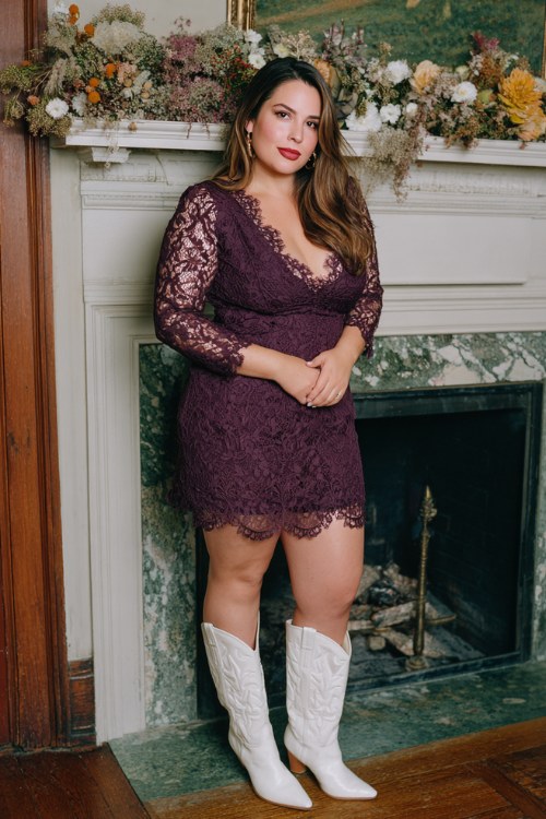 A plus size woman wears a plum lace cocktail dress with scalloped edges, styled with white cowboy boots
