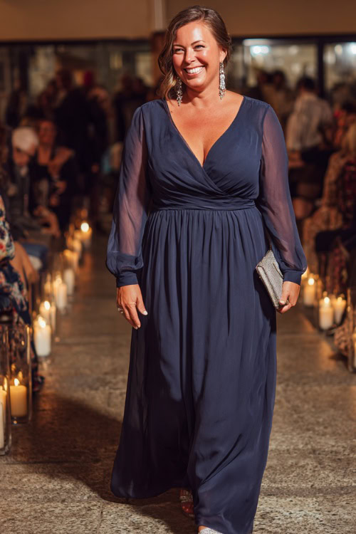 A plus-size woman wears a navy blue chiffon A-line gown with long sleeves and a flowy skirt, accessorized with silver drop earrings and metallic sandals