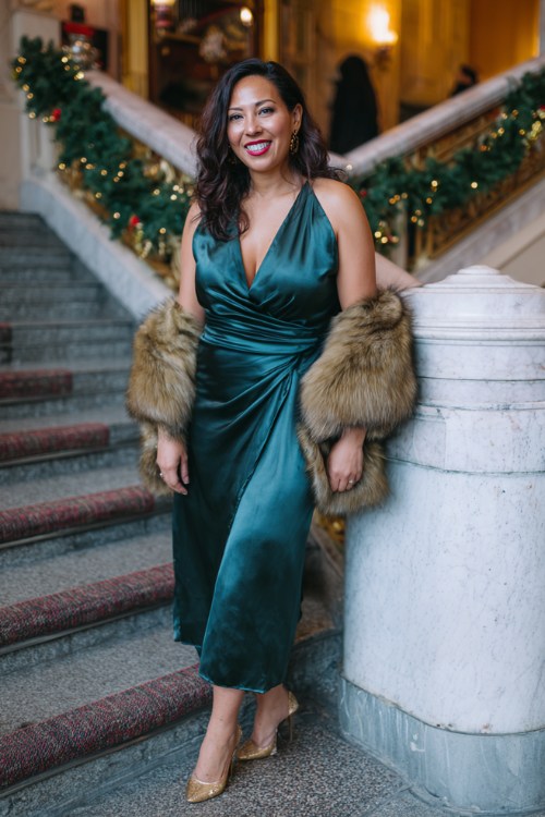 A curvy woman over 40 wears a deep emerald satin dress with a wrap neckline, fur shawl