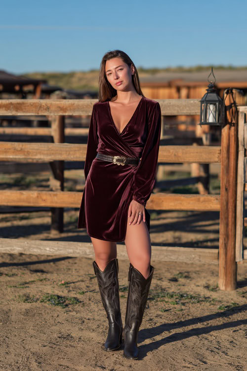 A woman wears a maroon velvet wrap dress with long sleeves, paired with tall black cowgirl boots and a statement belt