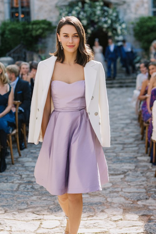 A woman wears a lilac A-line dress with a belted waist, paired with a white linen jacket