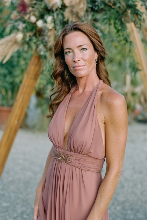 a woman over 50 in a dusty rose chiffon cocktail dress with beaded waistline, standing near a simple wooden ceremony arch