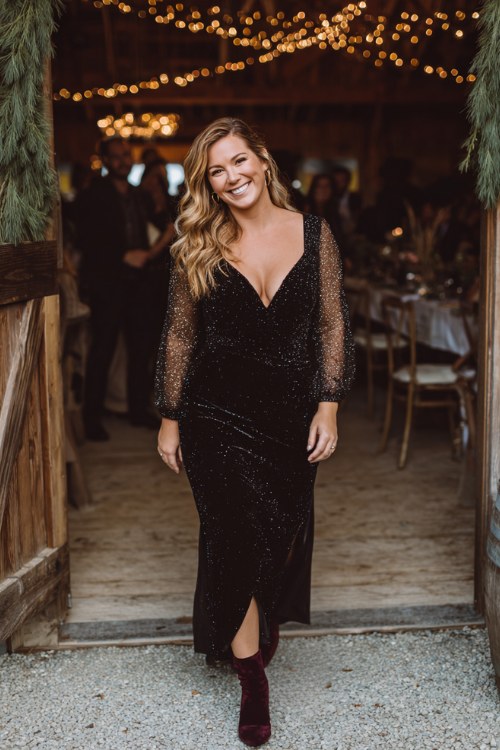 A curvy woman wears a black sequin cocktail dress with sheer mesh sleeves and dark plum velvet boots 2